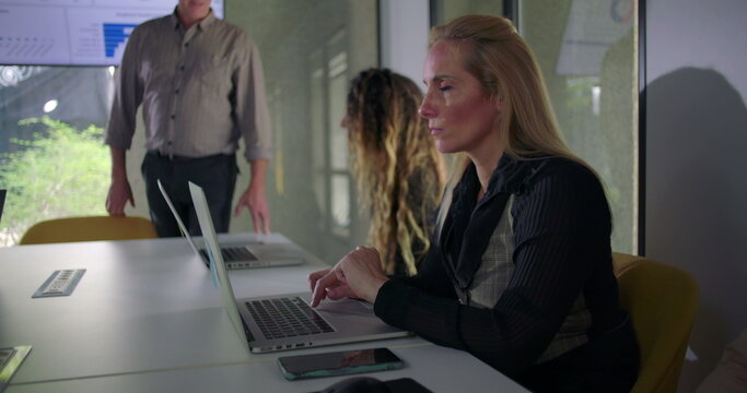 Blonde businesswoman working on laptop with serious expression during group meeting in modern office while coworkers and team leader collaborate nearby - Powered by Adobe