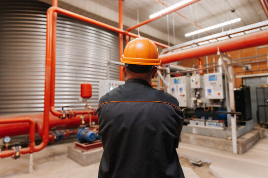 Engineer in hard hat inspecting industrial pump room with red pipes and control panels