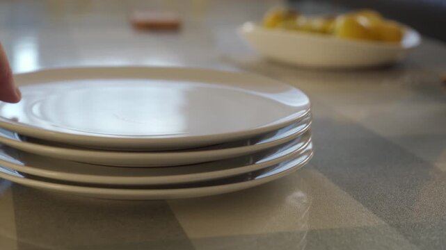 A stack of ceramic plates is set down by a hand onto a table with a plastic tablecloth. The hard plates hitting the surface creates a noticeable thud.