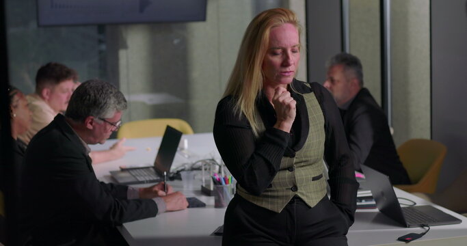 Blonde professional woman appearing tense and deep in thought during team meeting in office, standing near table as coworkers focus on work - Powered by Adobe