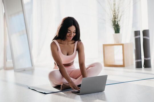 A fit young black woman sits in a lotus pose on a mat at home, using her laptop to communicate with her trainer. She searches for new workout videos during her fitness routine. - Powered by Adobe