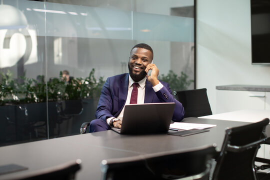 Positive young black businessman working on laptop at office, having phone conversation with his personal assistant or business partner, looking at camera and smiling, copy space - Powered by Adobe