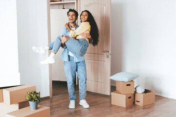 A couple is having fun moving into their new home. The man playfully lifts the woman while surrounded by unpacked boxes and bright sunlight. Happiness fills the room.