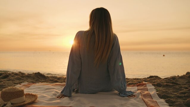 A woman enjoys the tranquil sunset, sitting on a blanket with golden sand beneath her and gentle waves in the background, feeling the evening breeze.