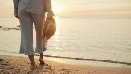 Walking along a serene beach at sunset while holding a straw hat, enjoying the tranquility of nature and the gentle waves