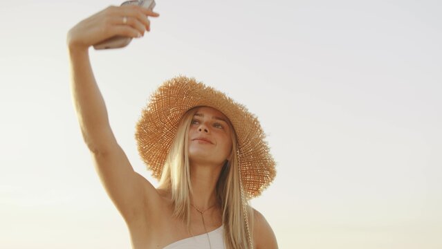 Young woman enjoys golden hour at beach while taking a selfie, wearing a stylish straw hat, capturing the moment filled with joy and relaxation - Powered by Adobe