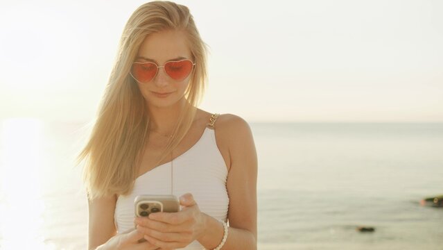 Young woman enjoys summer sunset at the beach while using her smartphone with heart-shaped sunglasses reflecting the golden light