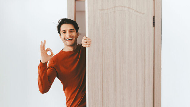 A young man stands in front of an open door, smiling and making an okay gesture with his hand. The room has a warm, inviting atmosphere with light walls and wood accents.