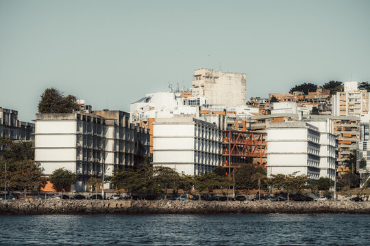 Telephoto view of coastal Niteroi buildings with geometric white blocks, dense hillside housing, trees and cars along a rocky waterfront under clear daylight