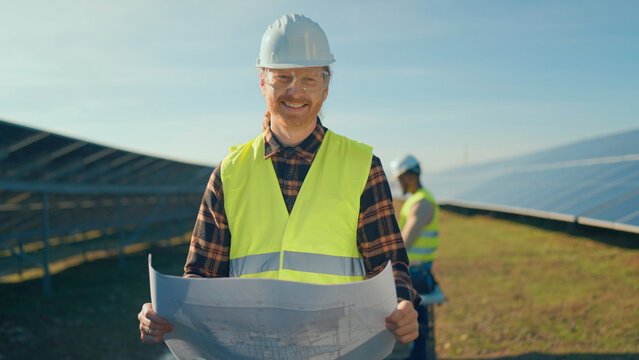Engineer holds blueprints while overseeing solar panel installation at a renewable energy site in sunny weather