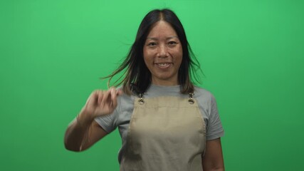 Woman showing peace sign hand and smiling in a green studio while wearing beige apron and gray tshirt, short black hair; playful confidence. - Powered by Adobe