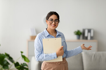 A cheerful arab female psychologist stands in an office, holding a clipboard and gesturing with her...