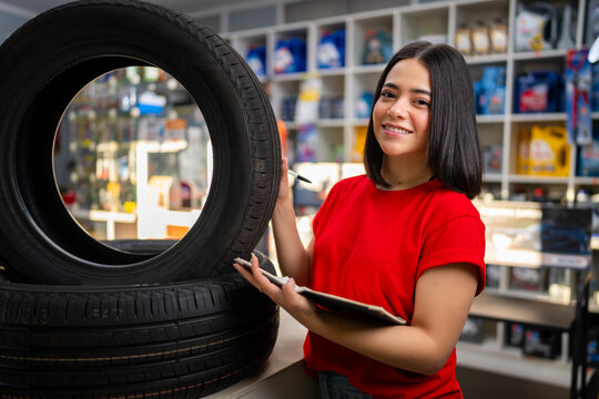 Young woman mechanic smiling, checking inventory of tires and auto parts in a garage store
