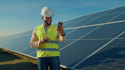Man in safety vest and hard hat smiles while checking phone near solar panels under clear sky
