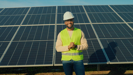 Smiling worker in safety vest stands proudly in front of solar panels during sunny day at renewable...
