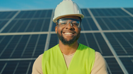 Professional smiling worker in safety gear stands confidently at solar energy farm during bright sunny day