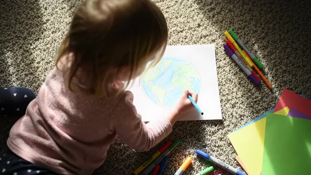 High-angle time-lapse video of a child drawing Earth with a blue pencil on beige carpet with art supplies, golden hour light. Creativity & environmental awareness.
