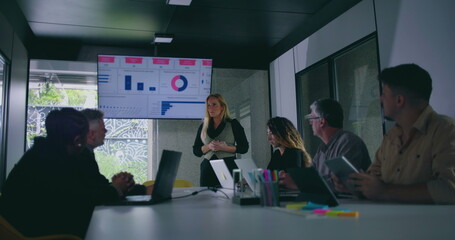 Businesswoman addressing team beside chart screen while colleagues observe during corporate meeting in brightly lit conference room