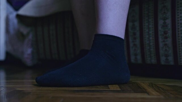 Experience the quiet intimacy of a person's foot, clad in a black sock, gently tapping on a polished wood floor beside a bed in a dimly lit bedroom.