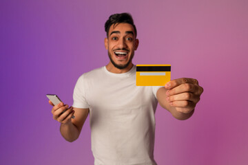 Happy millennial Arab man holds his cellphone in one hand and a credit card in the other, celebrating remote shopping. The neon lights create a vibrant background for his joyful moment.