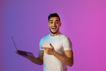 Young Arab man is happily pointing at his laptop while working online. The setting features lively neon lights, creating a modern and energetic atmosphere for his remote meeting.