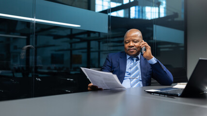 African American businessman reviewing documents while talking on cellphone in modern office. Focused mature executive handling work tasks with confidence and clarity