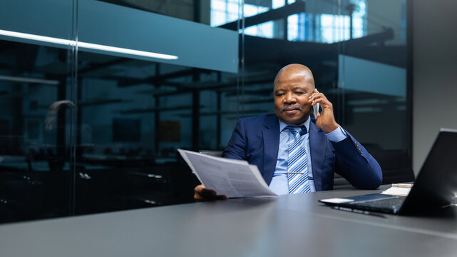 African American businessman reviewing documents while talking on cellphone in modern office. Focused mature executive handling work tasks with confidence and clarity