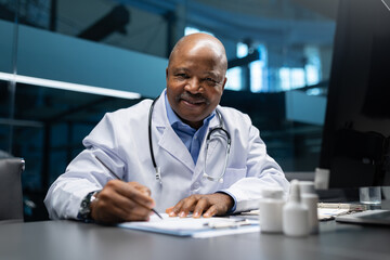 Smiling mature African American doctor writing notes at his desk. Confident medical professional working on patient information in a modern clinical office environment