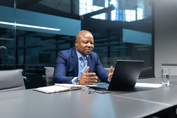 Mature African American businessman having an online meeting on his laptop, making video call, speaking and presenting confidently in modern office conference room.