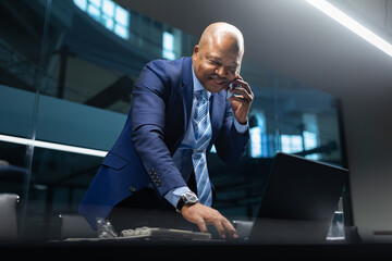 Mature African American businessman standing and talking on the phone while working on his laptop, representing dynamic leadership and multitasking in a modern office