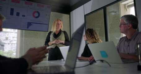 Businesswoman explaining data during meeting as team listens attentively in modern office with laptops and digital charts on display screen