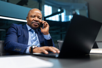 Mature African American businessman smiling while talking on the phone at his office desk. Confident corporate executive multitasking with laptop and documents