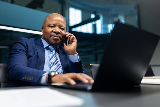 Mature African American businessman smiling while talking on the phone at his office desk. Confident corporate executive multitasking with laptop and documents - Powered by Adobe