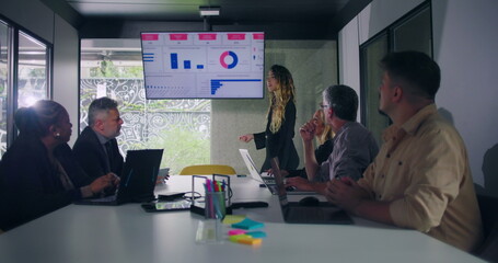 Businesswoman explains data chart to engaged team during office presentation while group of professionals seated around conference table watch screen