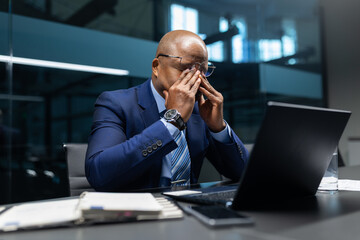 Mature african american businessman rubbing his eyes in front of a laptop, showing fatigue and stress while working long hours in a modern corporate office environment