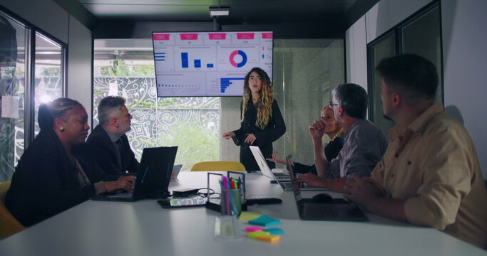 Businesswoman explains data chart to engaged team during office presentation while group of professionals seated around conference table watch screen - Powered by Adobe