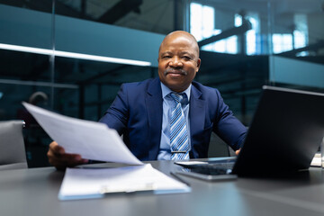 Confident mature African American businessman sitting at desk in modern office, reviewing documents with a friendly smile. Professional corporate atmosphere and leadership mood