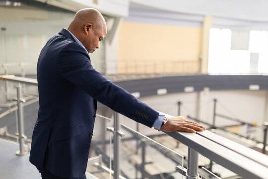 Mature African American businessman standing by railing, head lowered in stress or deep thought. Executive facing workplace pressure in a modern architectural environment