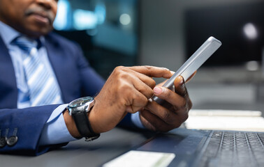 Closeup of mature African American businessman using his smartphone at the office desk. Focused executive checking messages or data during work, showcasing modern corporate workflow