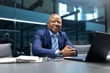 A mature African American businessman sitting confidently at his desk, smiling while posing in a modern office setting, representing leadership, professionalism, and success