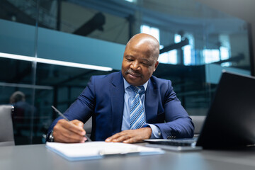 Mature African American businessman writing notes at his desk in a modern office setting. Focused executive working on paperwork with a calm, professional expression.