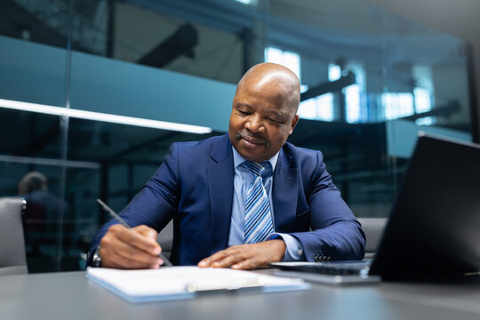 Mature African American businessman writing notes at his desk in a modern office setting. Focused executive working on paperwork with a calm, professional expression. - Powered by Adobe
