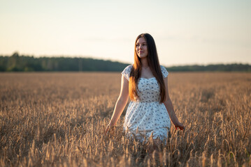 Girl in a light dress standing in a wheat field at sunset. A young woman in a white dress standing among golden wheat ears, looking into the distance, harmony, calmness, connection to nature