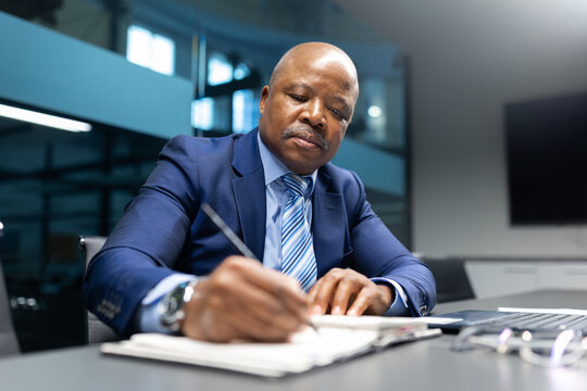 Mature African American businessman writing in his notebook during a work session in office, focused on documentation and planning in a modern office environment - Powered by Adobe