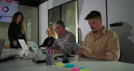 Business meeting with diverse team focused on digital presentation, as male employee on right uses tablet while others engage with charts and laptops around conference table