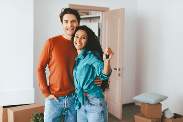 A happy couple stands together, smiling while holding up a set of house keys. They are in a room with moving boxes, indicating they just moved into a new home.