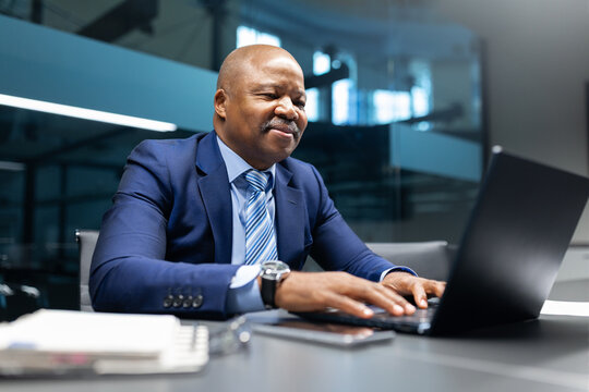 Mature smiling African American businessman working on his laptop in bright office, focused on completing tasks and managing responsibilities in a professional corporate environment