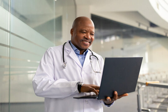 Mature African American doctor using a laptop in a modern building interior. Friendly medical professional reviewing patient data and working digitally with confidence and precision
