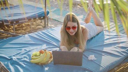 Young woman lounges on beach bed using laptop while enjoying fresh fruits under sunlight