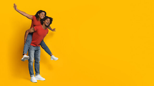 A joyful couple poses against a vivid yellow backdrop, with one partner giving the other a playful piggyback ride. Both wear casual red shirts and display cheerful expressions.